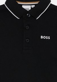 Čierna polo košeľa vyrobená z textúrovanej látky, s klasickým golierom, tromi gombíkmi a bielym logom "BOSS" na ľavej hrudi, s kontrastnými akcentami goliera.