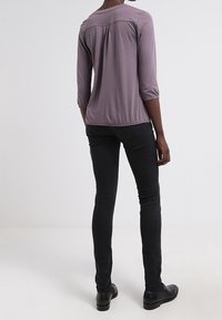 Maglietta grigia a maniche a tre quarti con dettaglio arricciato sul retro, abbinata a pantaloni neri aderenti e scarpe scure. Tessuto dalla texture liscia.