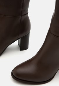 Stivaletti in pelle marrone con punta affusolata e texture liscia. Presentano un tacco spesso e dettagli di cucitura minimi.