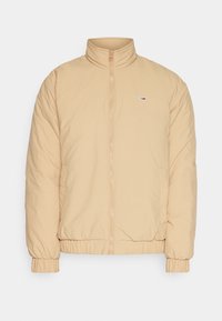 Beige prošívaná bunda s vysokým límcem, zipovým zapínáním, elastickými manžetami a postranními kapsami, s malou nášivkou loga na hrudi.