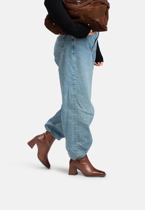 Person trägt hellblaue, weite Jeans und braune, absatzstarke Stiefeletten und hält eine große braune Wildledertasche unter einem Arm.