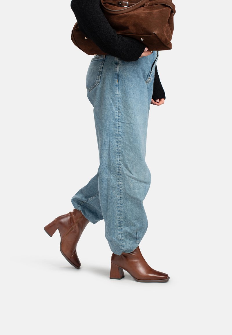 Persona vistiendo jeans holgados azul claro y botas tobilleras marrones con tacón, llevando un bolso grande de ante marrón bajo un brazo.