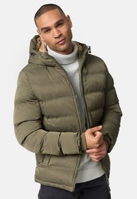 Oliven grøn pufferjakke med hætte, der har lynlås foran, ribbede manchetter og sidelommer. Båret over en grå strikket turtleneck.