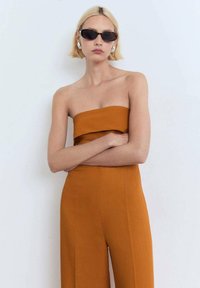 Strapless, aansluitend top in gebrande oranje, gecombineerd met wijde broek in dezelfde kleur. Model draagt donkere zonnebril en metalen oorbellen.