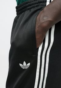 Μαύρο αθλητικό παντελόνι με ελαστική μέση, διακοσμημένο με τρεις λευκές πλευρικές ρίγες και μικρό γκρι λογότυπο Adidas trefoil. Ομαλή υφή.