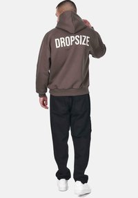Brun hættejakke med stor hvid tekst "DROPSIZE" på ryggen, parret med sorte bukser og hvide sneakers. Har hætte og ribbede manchetter.