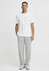 T-shirt blanc à manches courtes avec un petit emblème sur la poitrine, associé à un pantalon de survêtement gris clair et des baskets grises, debout devant un fond uni.