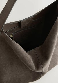 Geantă tote din suede maro, cu o textură moale, o deschidere mare, un buzunar intern cu fermoar și un design minimal. Detalii cusute pe margini.
