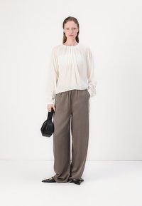 Valkoinen laskostettu pitkähihais blouse, joka on yhdistetty löysiin taupe-housuihin ja mustiin kärkitossuihin. Malli pitää pieniä mustaa käsilaukkua.