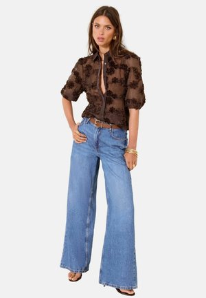 Woman wearing a sheer brown blouse with floral patterns, wide-leg blue jeans, a brown belt, gold bracelets, and black open-toe heels.