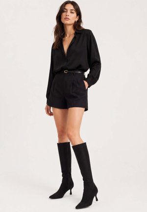 Frau trägt schwarze Langarmbluse, hoch taillierte schwarze Shorts mit Gürtel und schwarze kniehohe Stiefel mit Absatz, steht vor einfarbigem Hintergrund.