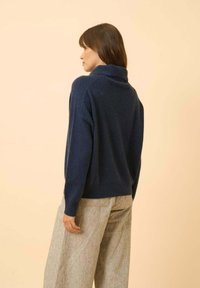 Námořnická modrá pletená svetr s vysokým límcem, měkkou texturou a volným střihem, doplněný o světle šedé široké kalhoty s jemným vzorem.