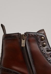 Bottine en cuir marron avec des détails décoratifs de brogue, une fermeture éclair latérale et des accents en métal. Texture lisse et design robuste.