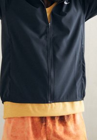 Person wearing a navy zip-up jacket, mustard yellow shirt underneath, and orange tie-dye pants, standing against a plain background.