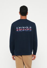 Moški v temno modri trenerki z večbarvnim napisom "1985 Tommy Jeans Established" na hrbtni strani, v kombinaciji s svetlimi hlačami, stoji ob belem ozadju.