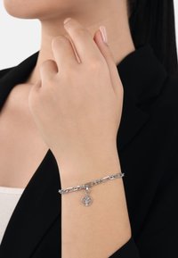 Bracciale in argento con anelli ovali e un ciondolo a forma di albero circolare. Texture liscia, indossato su un polso, abbinato a un blazer nero.