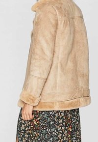 Giacca in shearling marrone chiaro con bordo di pelliccia sui polsini e sull'orlo, indossata su una gonna nera con motivo floreale e fiori multicolori di piccole dimensioni.