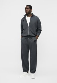 Homme debout les mains dans les poches, portant une veste zippée gris foncé, un pantalon assorti, un t-shirt blanc et des baskets blanches sur un fond uni.