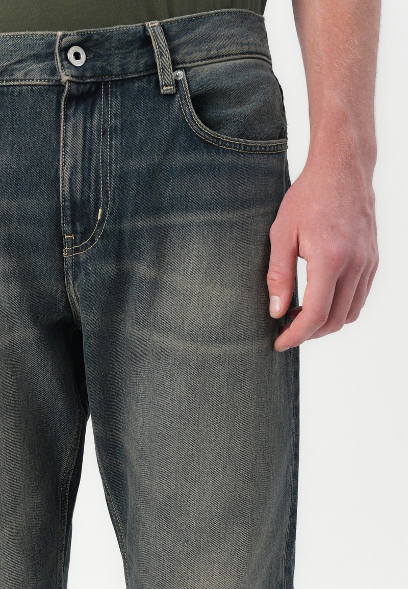 Dark blue denim jeans with a faded finish, five-pocket design, button closure, and contrast stitching. Hand resting on the left thigh.