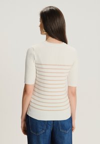 Mujer con cabello largo y castaño lleva una blusa de punto blanco con rayas horizontales beige y pantalones de mezclilla azules, de espaldas a la cámara.