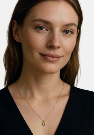 Junge Frau mit hellbraunen Haaren, die ein schwarzes Oberteil und eine goldene Halskette mit einem runden Anhänger trägt, auf dem der Buchstabe "G" eingraviert ist.