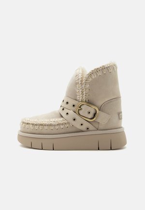 Beige suede ankle boot with thick rubber sole, stitched edges, adjustable strap, and buckle detail, lined with soft shearling interior.
