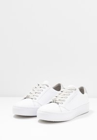 White low-top sneakers with thick soles, beige laces, and silver detail on the heel, featuring the "Tommy Hilfiger" logo on the side.