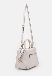 Jasnobeżowa skórzana torba na ramię z złotym zamkiem, pierścieniami, podwójnymi uchwytami u góry oraz długim regulowanym paskiem na białym tle.