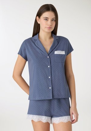 Giovane donna che indossa una maglietta blu a maniche corte con bottoni e pantaloncini abbinati con pois bianchi e rifiniture in pizzo sui pantaloncini e sulla tasca.