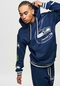 Felpa con cappuccio blu navy con il simbolo dei Seattle Seahawks, dotata di cordini bianchi e dettagli verdi sulle maniche. Tessuto morbido.