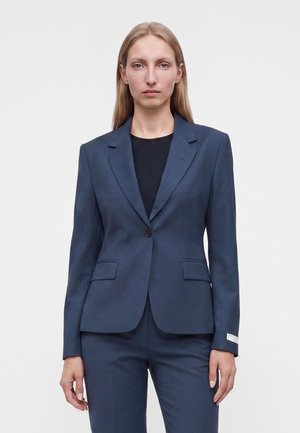 Femme aux cheveux blonds raides porte un blazer bleu foncé ajusté et un pantalon assorti par-dessus un haut noir, debout devant un fond uni.