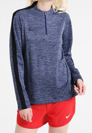 Kobieta ubrana w niebieską bluzę z długim rękawem i zamkiem do połowy szyi, ozdobioną czarnymi paskami oraz czerwone sportowe spodenki Nike.