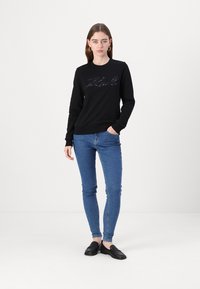 Felpa nera con scollo rotondo e dettaglio testuale ricamato, abbinata a jeans blu slim-fit e scarpe slip-on nere.