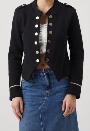 Schwarze Militärjacke mit goldenen Knöpfen, strukturiertem Kragen und goldenen Akzenten an den Manschetten, kombiniert mit einem strukturierten weißen Oberteil und einem blauen Denim-Rock.