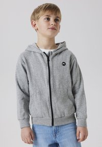 Ragazzo giovane che indossa una felpa grigia con zip e jeans blu, in piedi leggermente girato con le mani rilassate ai lati.