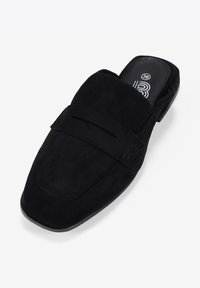 Zapato negro de ante sin cordones con punta cuadrada, suela plana y correa decorativa en la parte superior. Presenta detalles de costura sutiles.