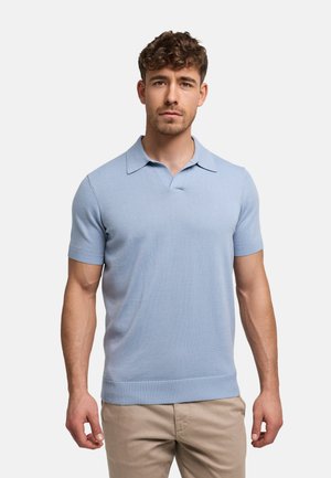 Man met kort krullend haar, gekleed in een lichtblauw poloshirt met korte mouwen en beige broek, staand tegen een effen lichte achtergrond.