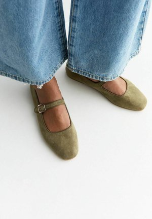 Voeten in olijfgroene suède Mary Jane flats met gespbandjes en wijd uitlopende blauwe jeans op een witte achtergrond.