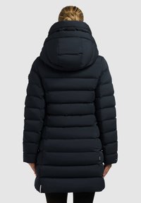 Mujer vestida con un largo abrigo acolchado negro con una gran capucha, vista desde atrás sobre un fondo liso.