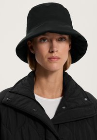 Kvinna som bär en svart bucket hat och en kviltad svart jacka över en vit ribbad topp, med en neutral uttryck och beige bakgrund.