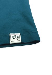 Teal cotton t-shirt corner with a white label featuring black crossed utensils and initials "B R." Seam details are visible.