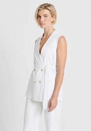 Blonde Frau trägt ärmellosen weißen Blazer mit Gürtel und doppelter Knopfleiste sowie passende weiße Hose, steht vor einfachem Hintergrund.