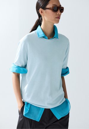 Femme en lunettes de soleil portant un pull bleu clair par-dessus une chemise bleu vif à col avec les manches retroussées et un pantalon noir, les mains dans les poches.