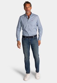 Blauw gemêleerd overhemd met lange mouwen, dichtgeknoopt bij de kraag, gecombineerd met lichtblauwe jeans en witte sneakers. Bevat een subtiel logo op de borst.
