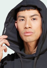 Sudadera negra con un logo visible, hecha de material suave, con capucha ajustable y un corte relajado, parcialmente recogida que revela una camiseta azul por debajo.