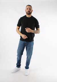 Maglietta nera a maniche corte, jeans in denim blu e sneakers bianche. Braccio tatuato visibile, in piedi contro uno sfondo bianco.