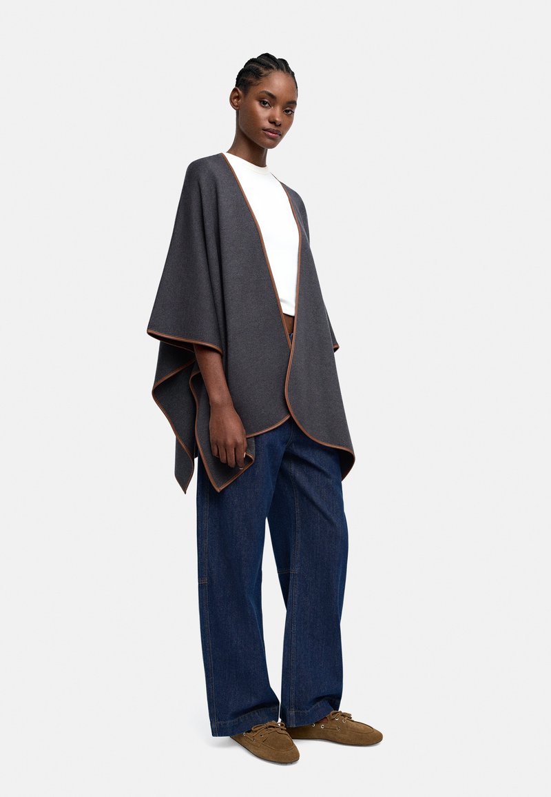 Ciemnoszara narzutka z pomarańczowymi akcentami, wykonana z przytulnego materiału. Posiada luźny, kanciasty krój, w zestawieniu z szerokimi dżinsami i brązowym obuwiem.