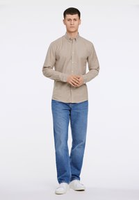 Camicia button-up marrone chiaro con maniche lunghe, abbinata a jeans blu e sneakers bianche. La camicia ha un colletto e polsini con un bottone.