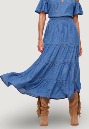 Mujer que lleva un vestido midi azul fluido con mangas cortas con volantes y botas marrones de ante con flecos, de pie contra un fondo liso.