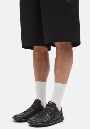 Chaussures en cuir noir avec des lacets élastiques, associées à des chaussettes blanches jusqu'à la cheville. Portées avec un short noir mi-long, affichant un look décontracté.